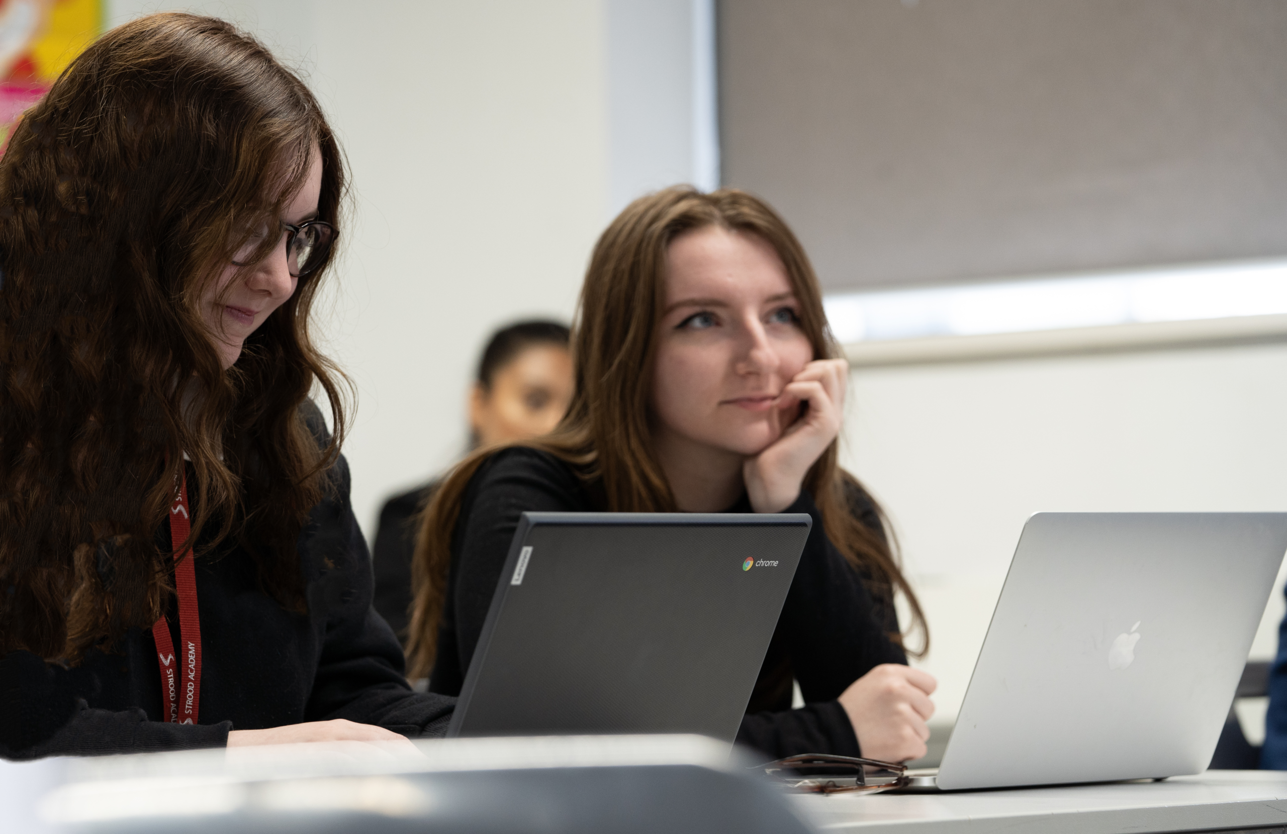 Two students working on their respective laptops listening to something in class.