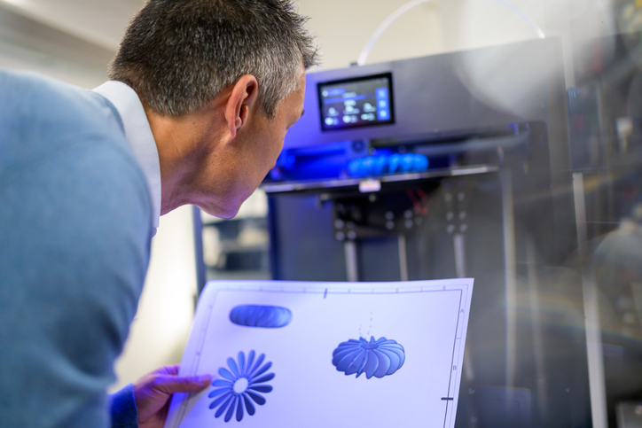 Back view of man holding technical drawing and observing 3D printing of part production. Producing of 3D object from digital model using additive manufacturing.