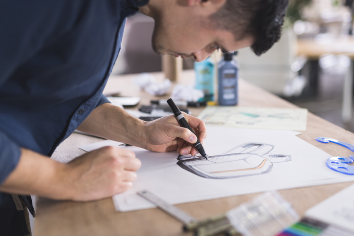 A graphic designer sketches out prototypes for an idea onto paper. He is left-handed and using a pen. There are various pens, papers, and art supplies setting on the desk. The shot is focused on the sketch and includes his hand and his head in profile as he leans over to concentrate.