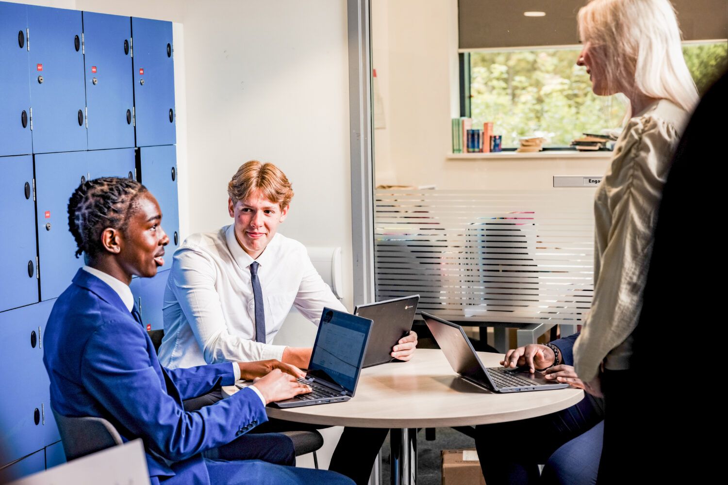 Two Sixth Form students using their Chromebooks in a classroom talking to a teacher