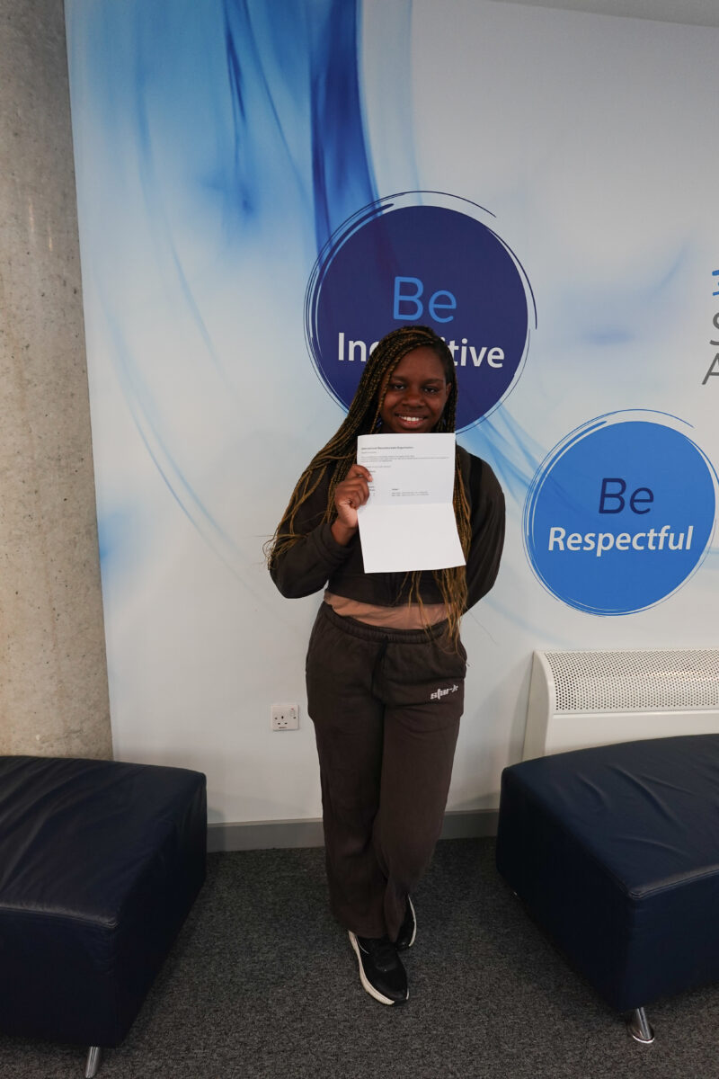 A young woman is smiling and holding up a document (likely her results) in front of her. She is standing in the same Leigh Academy Strood setting as the previous photo, with the "Be Inquisitive" and "Be Respectful" circles visible behind her.