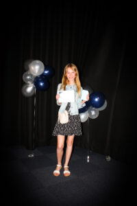 Young girl posing holding a certificate, in front of black backdrop with blue and silver balloons