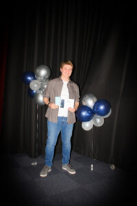 Young boy posing holding a certificate, in front of black backdrop with blue and silver balloons