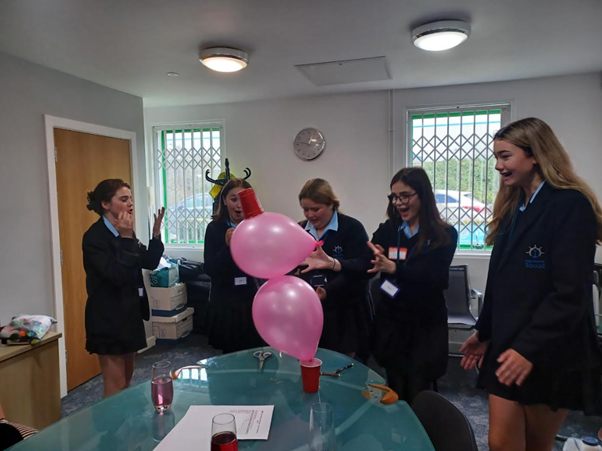 A group of students in school uniforms cheering and reacting with excitement as they balance pink balloons and red cups during an indoor activity.