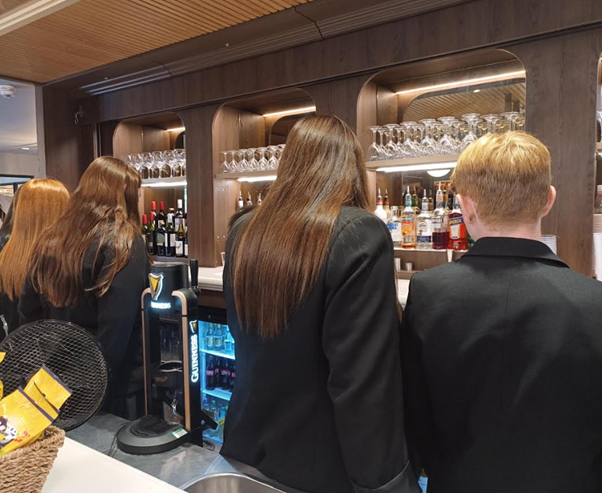 Four students in black blazers standing with their backs to the camera at a bar counter, which is stocked with various glasses and bottles.