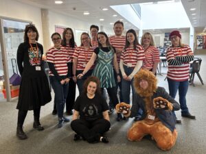 A group of adults in a spacious hallway dressed up in various costumes, including red and white striped shirts, a green dress, and a lion suit.