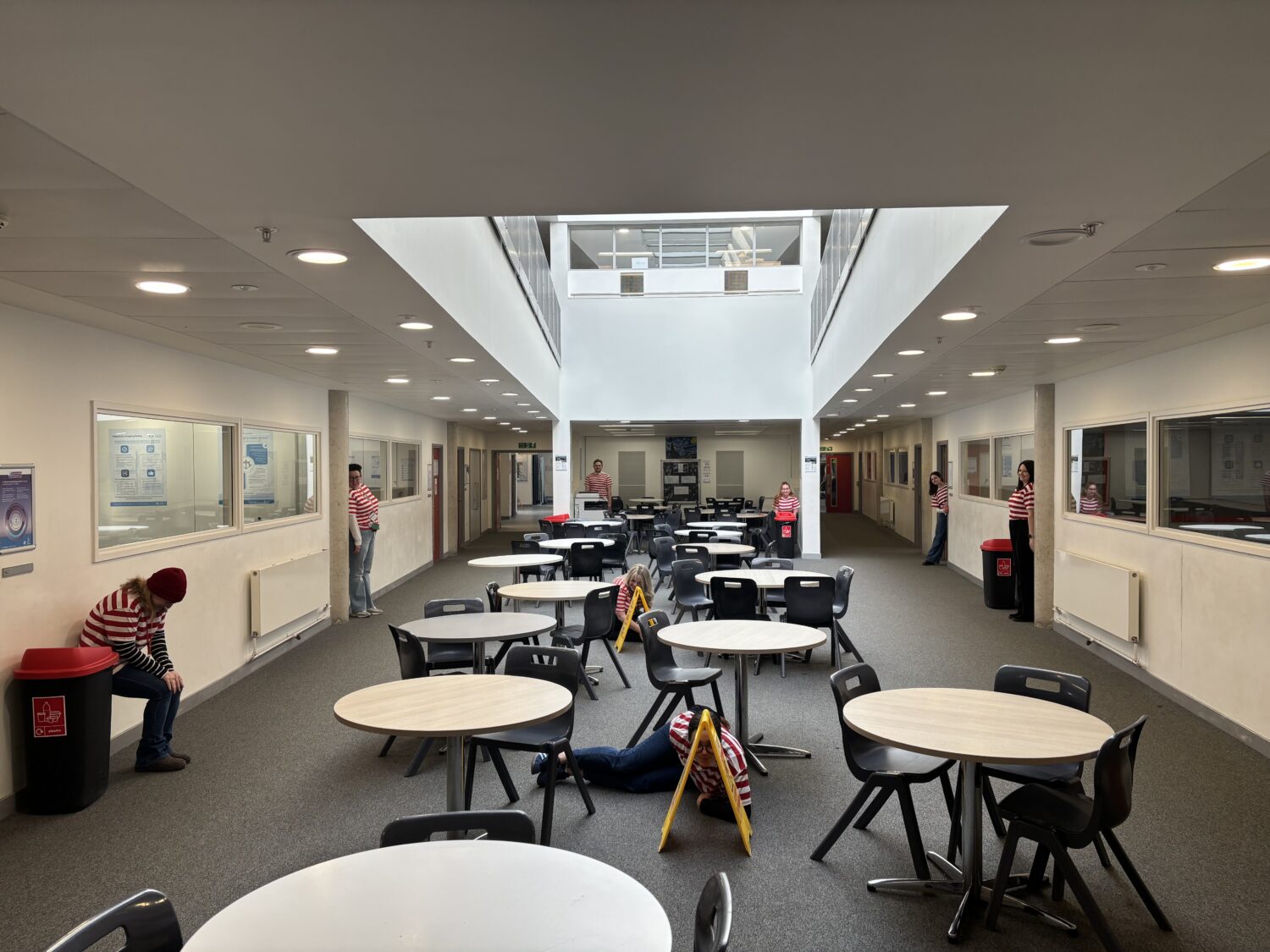 A wide view of a room with tables where several people in red and white striped shirts are playfully hiding behind bins, wet floor signs, and corners.