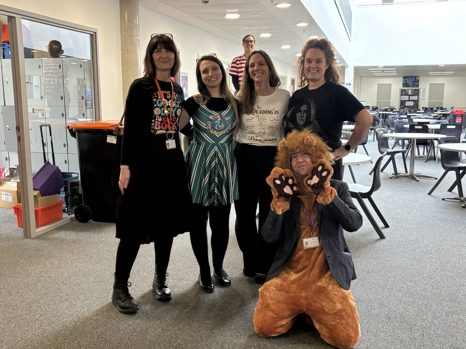 Five adults posing for a photo in a hallway wearing book-themed outfits, including a lion costume, with another person in a striped shirt peeking from the background.