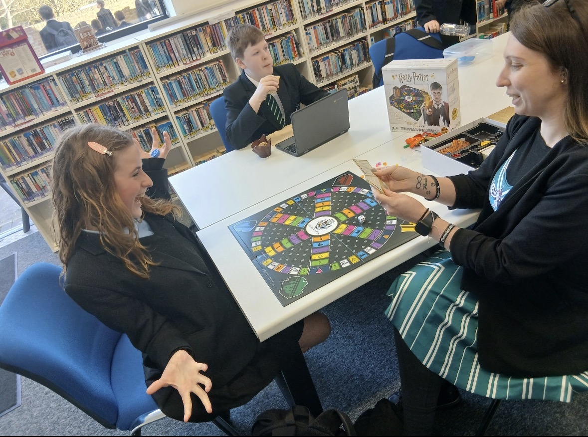 A teacher and two students excitedly playing a Harry Potter board game together at a table in a library.
