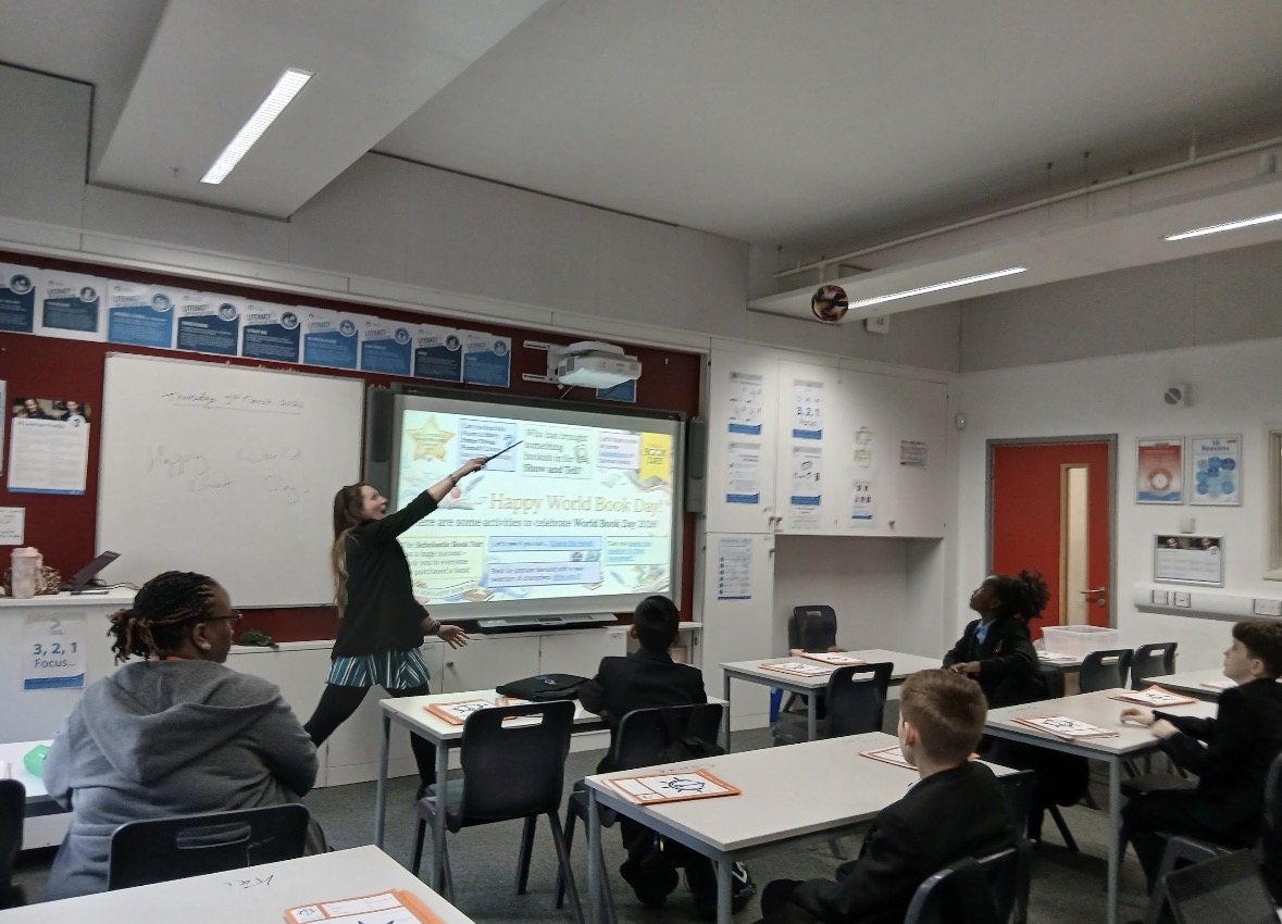 A teacher at the front of a classroom playfully tossing an object into the air next to a whiteboard displaying "Happy World Book Day!", as students watch from their desks