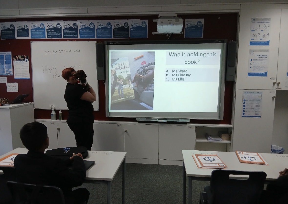 A teacher holding a small dog in front of a classroom whiteboard that shows a reading-themed quiz question, with students seated at desks in the foreground.