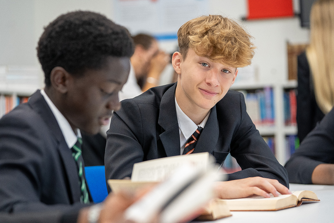 Two students reading books at a desk. One is looking at the camera smiling